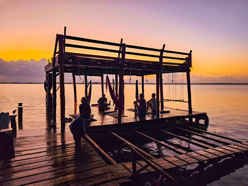 A sunrise yoga class at Casa Lamat. A small group of people sitting on yoga mats on a wooden dock on Bacalar Lagoon looking out at a bright orange sky.