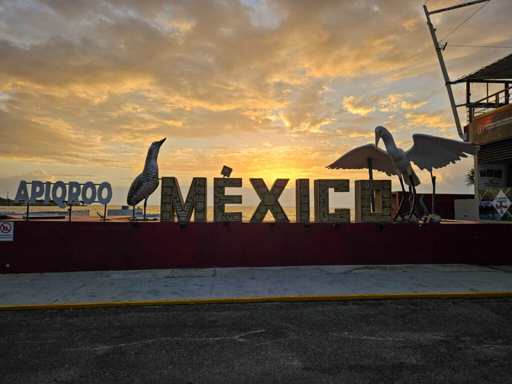 A decorated "Mexico" sign at Chetumal Ferry Port. Behind the sign, the sky is turning orange in the early morning sun.