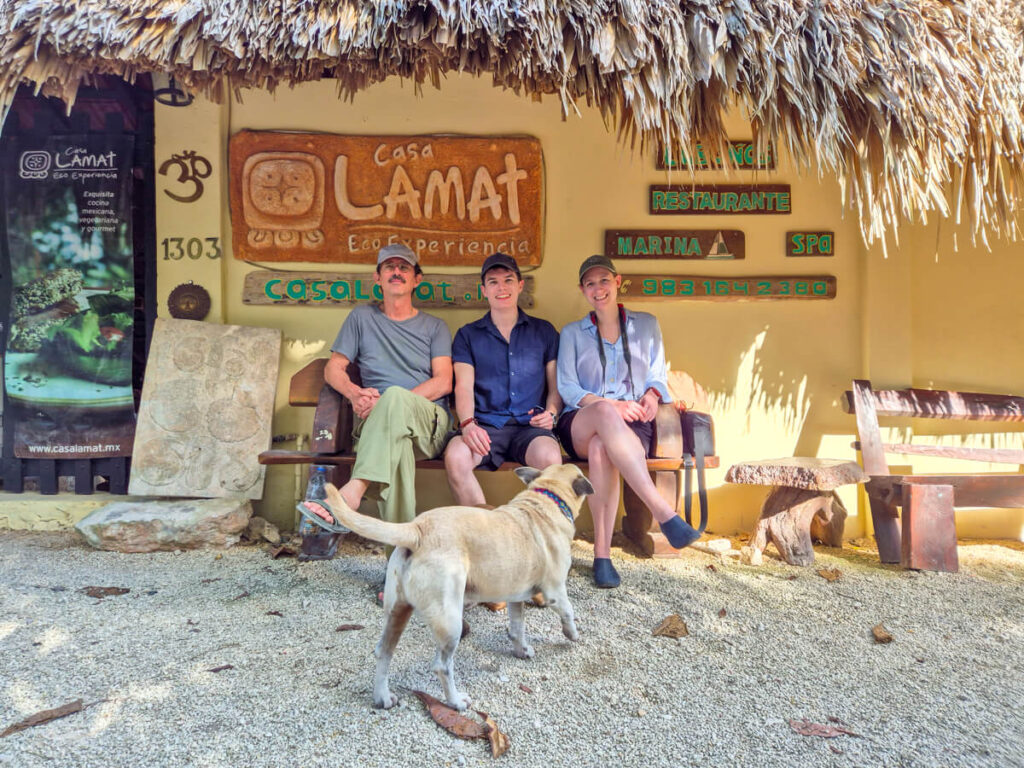 Dan and Lucy sitting smiling with Xavy, the founder of the Eco Hotel Casa Lamat in Bacalar. They are all sitting on a bench in front of a sign that reads "Casa Lamat Eco Experiencia".