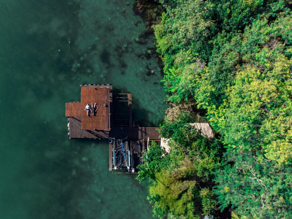 A section of Bacalar Lagoon as viewed from a drone. The water is a blue-green colour and matches well with the surrounding greenery. Lucy and Dan are lying down on a wooden dock looking up at the sky.