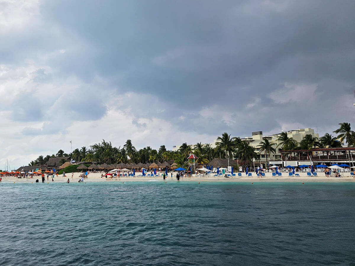 Isla Mujeres as viewed from a boat tour with Ocean Tours Mexico. The beach is lined with palapas, sun loungers and tourists.