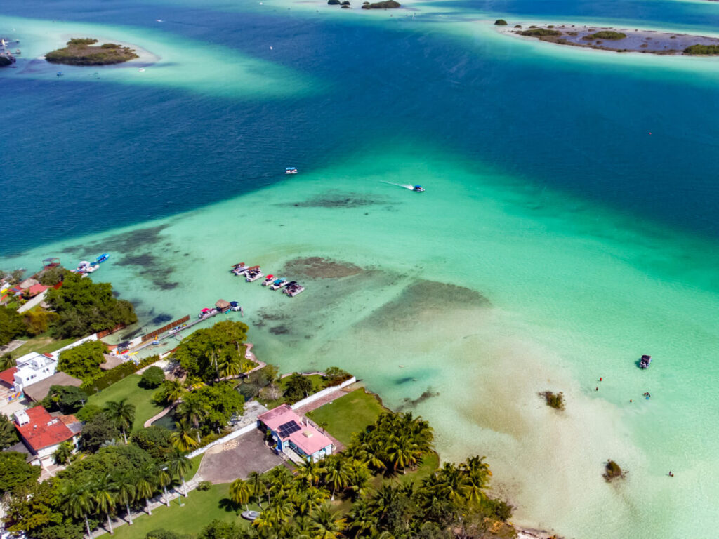 Boats and rafts moving slowly in the shallows of Bacalar Lagoon. Nearby, the water depth increases, resulting in a pattern of different shades of blue.