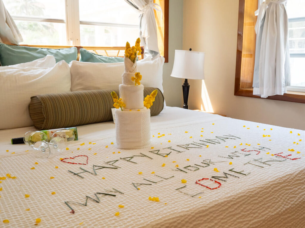 A birthday message on a hotel bed reads "Happy Birthday. May all your wish come true". Beside the message is a bottle of white wine and two glasses, as well as an arrangement of towels and flowers that looks like a cake.