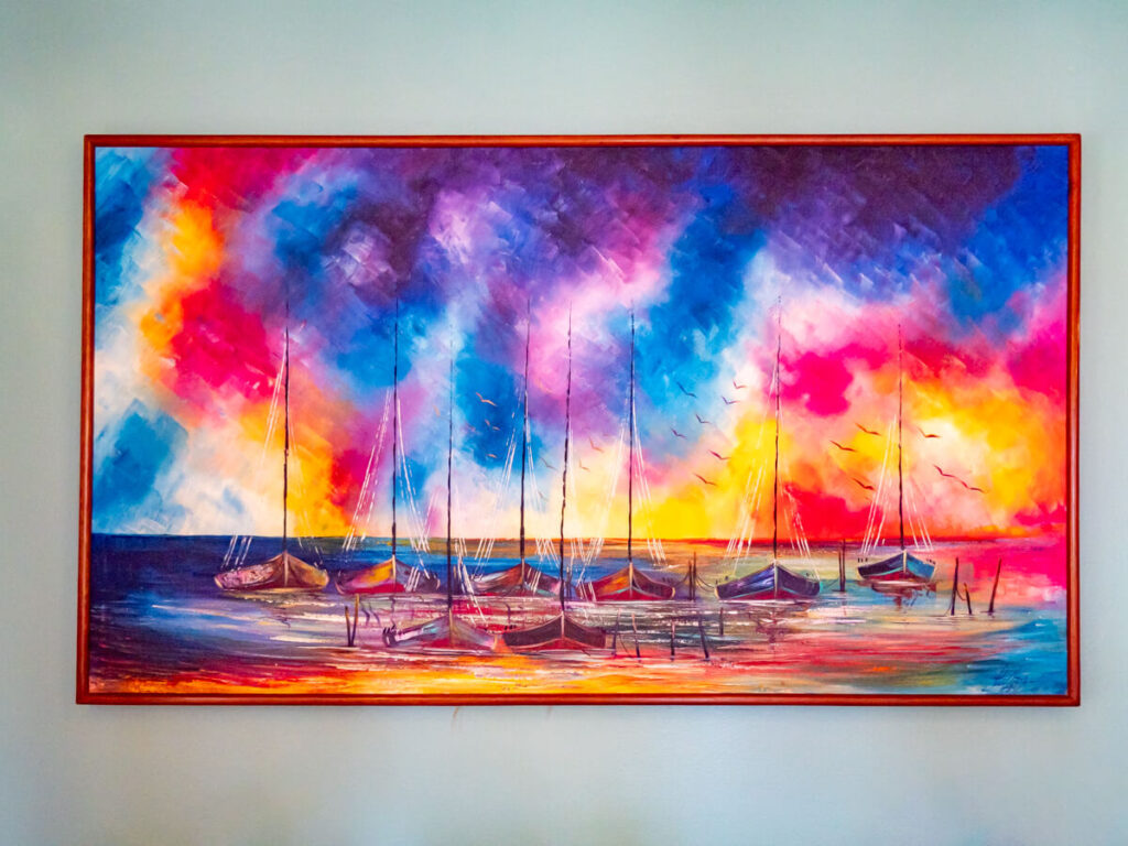 A painting of a bright, multi-coloured sky and a small group of sail boats moored in calm waters.