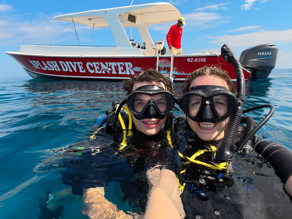 Lucy and Dan in SCUBA diving gear at the water's surface. They are smiling in front of a boat that has the text "Splash Dive Center" painted on its side.
