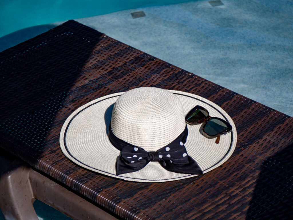 A large white holiday hat, decorated with a spotty bow, sitting on a sun lounger by a hotel pool. On top, there is a pair of brown sunglasses.