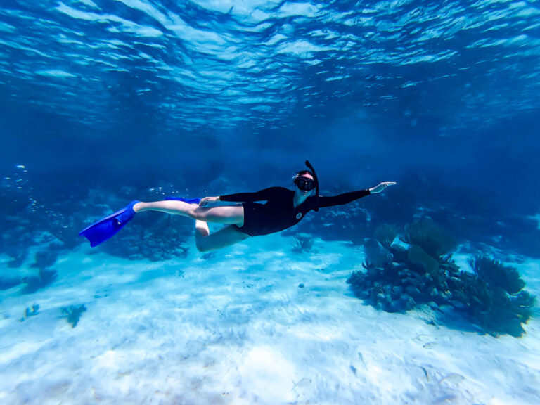 Lucy snorkelling in blue waters above white sand. She is extending her arm out and turned to face the camera.