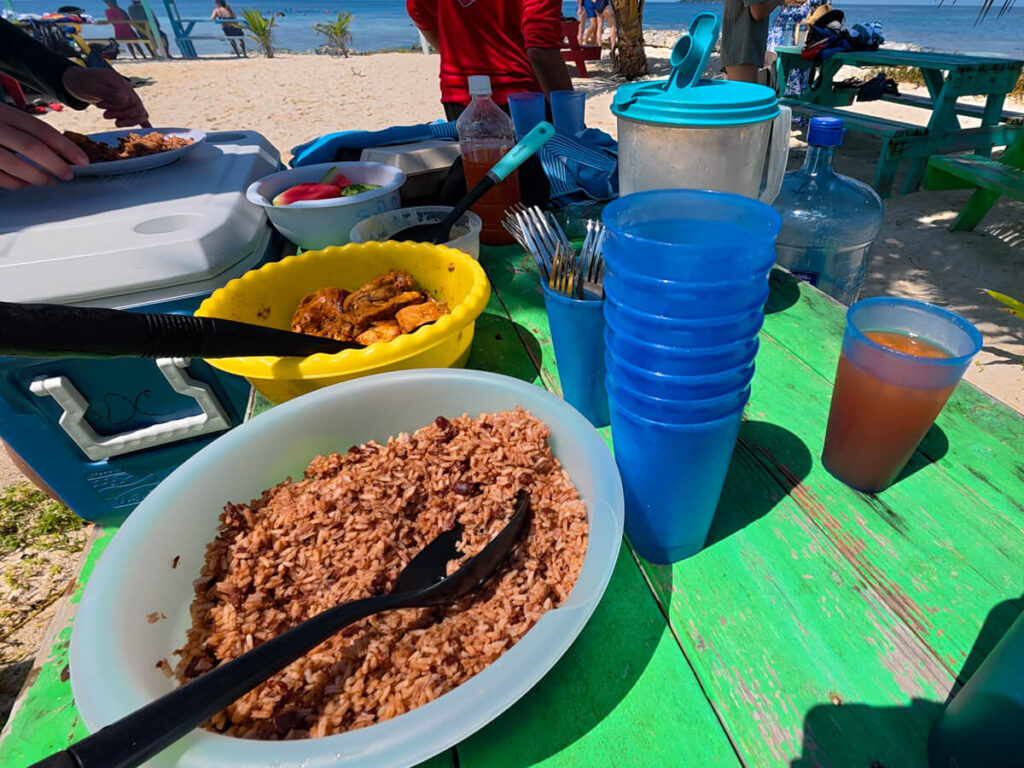 Food laid out for lunch on the beach. On a wooden table is fresh juice, rice and beans, BBQ chicken, potato salad and slices of watermelon.