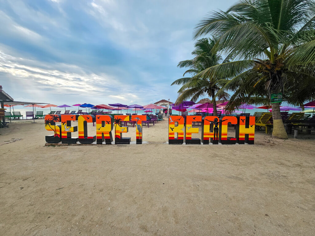 A colourfully-painted sign that reads "Secret Beach" against a backdrop of white sand, parasols, and the endless blue horizon.