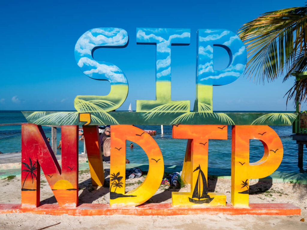 A sign for the Sip N' Dip beach bar on Caye Caulker against a backdrop of palm trees and calm seas.