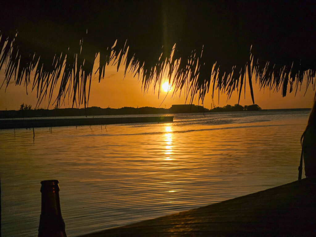 A bright orange sunset viewed from underneath a thatched palapa-style roof. This is the view from the Maggie's Sunset Kitchen, one of the best places to eat in Caye Caulker.