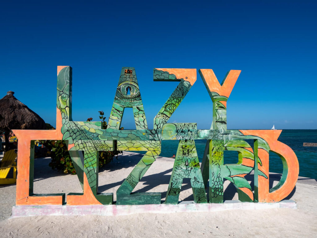 The Lazy Lizard sign in Caye Caulker. An image of a lizard is painted across the letters and the clear blue sky can be seen in the background.