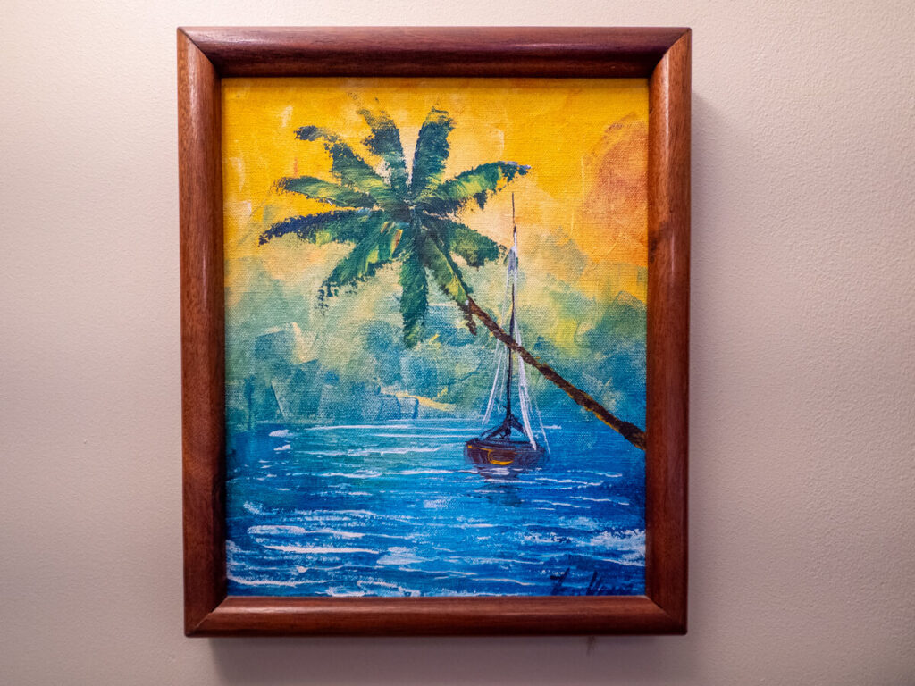 A painting of a lone sailboat on blue waters behind a tail palm tree.