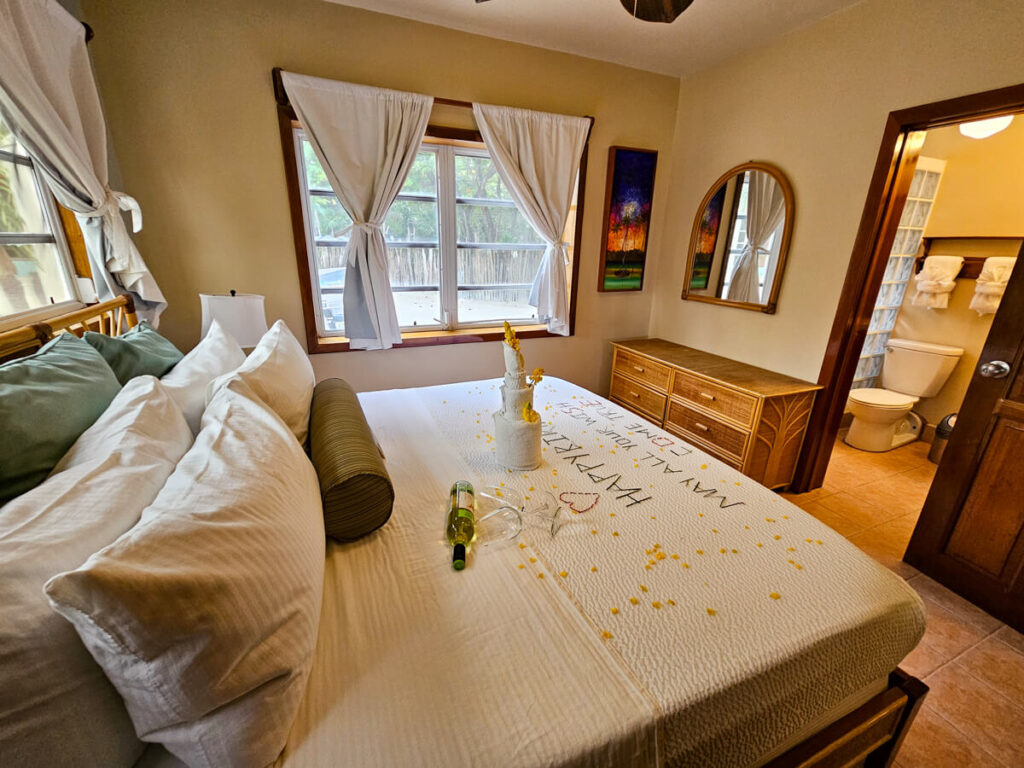 A bedroom in Belizean Shores Resort. Flower petals have been spread on the white bedding, and a bottle of wine has been placed on the bed as a birthday gift.