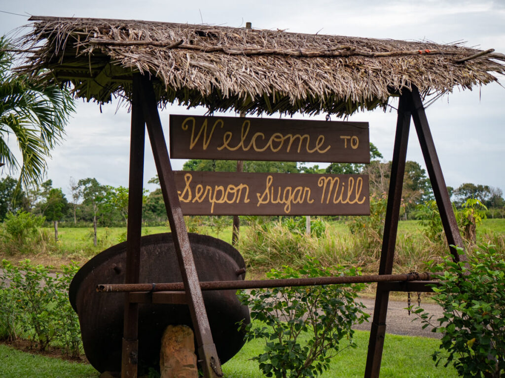 A sign that reads "Welcome to Serpon Sugar Mill" under the cover of a traditional palapa-style roof.
