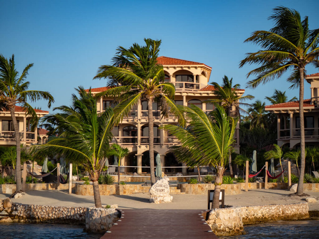 The front of Coco Beach Resort in San Pedro, Belize. The early morning light is casting a warm glow on the lavish, multi-storey buildings, with palm trees swaying in the wind. Coco Beach Resort is one of the most highly-rated hotels in San Pedro.