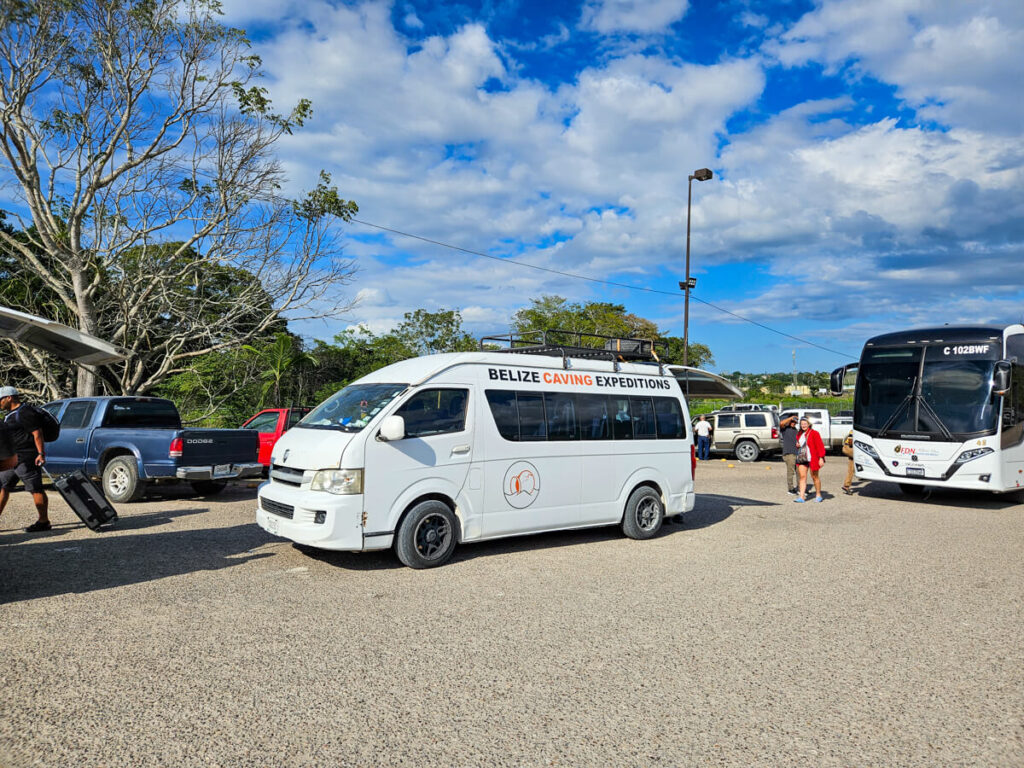 A tourist shuttle, parked in a busy car park, with the words "Belize Caving Expeditions" on the side. Taking a shuttle is one of the most affordable ways to get around Belize.