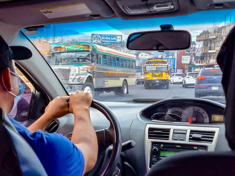 A driver navigating busy streets while driving in Guatemala. Colourful chicken buses can be seen out of the windshield.