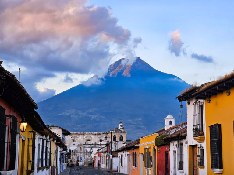 A volcano looming over a cobblestone street in Antigua, Guatemala, lined by colourful buildings.