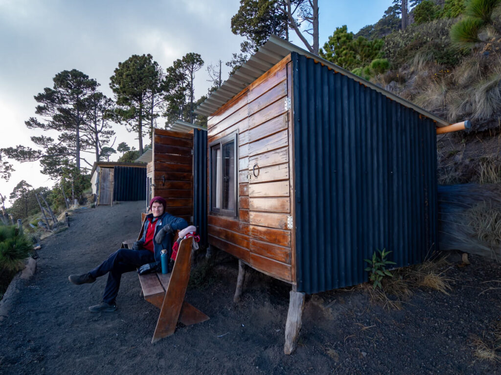 Dan smiling as he sits on a bench in warm clothing outside his wooden cabin at Acatenango Base Camp.
