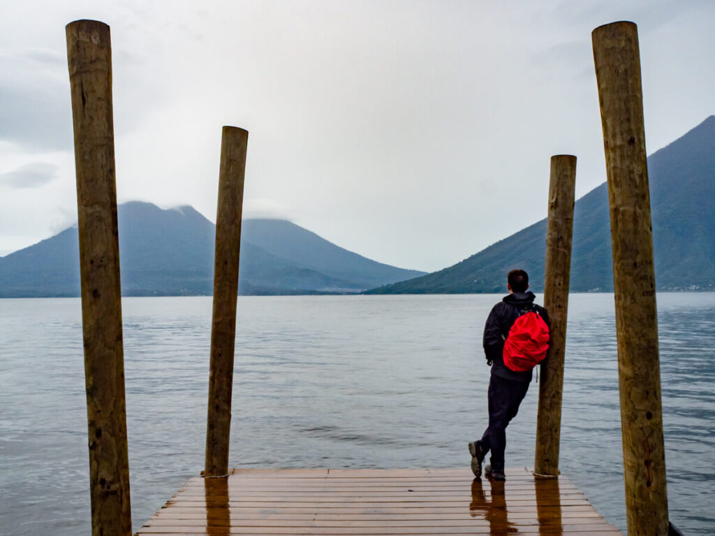 Dan in a black coat, wearing a rucksack with a red waterproof cover, leaning on a pole on a wooden dock, looking out at the volcanoes on Lake Atitlán.