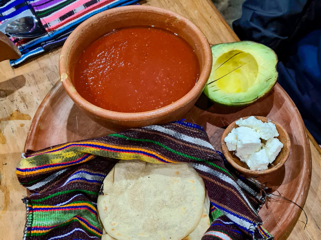 A bowl of red soup, tortillas, and half an avocado on a plate with a colourfully embroidered napkin.