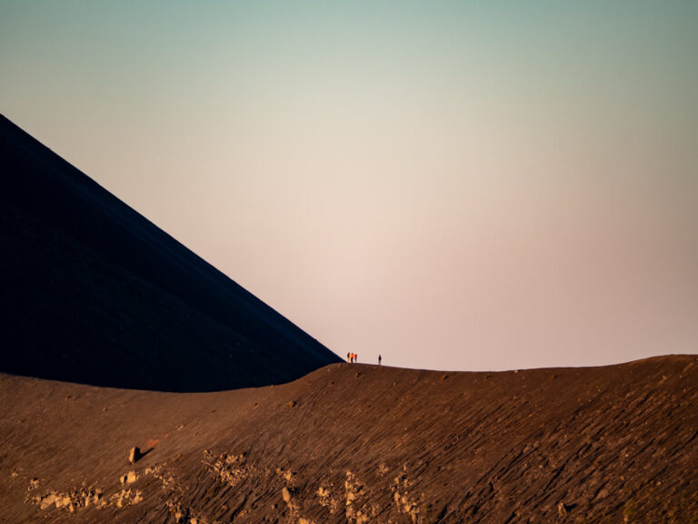 Hikers bathed in soft pink light at sunrise while standing on a ridge of the dark brown Fuego Volcano.