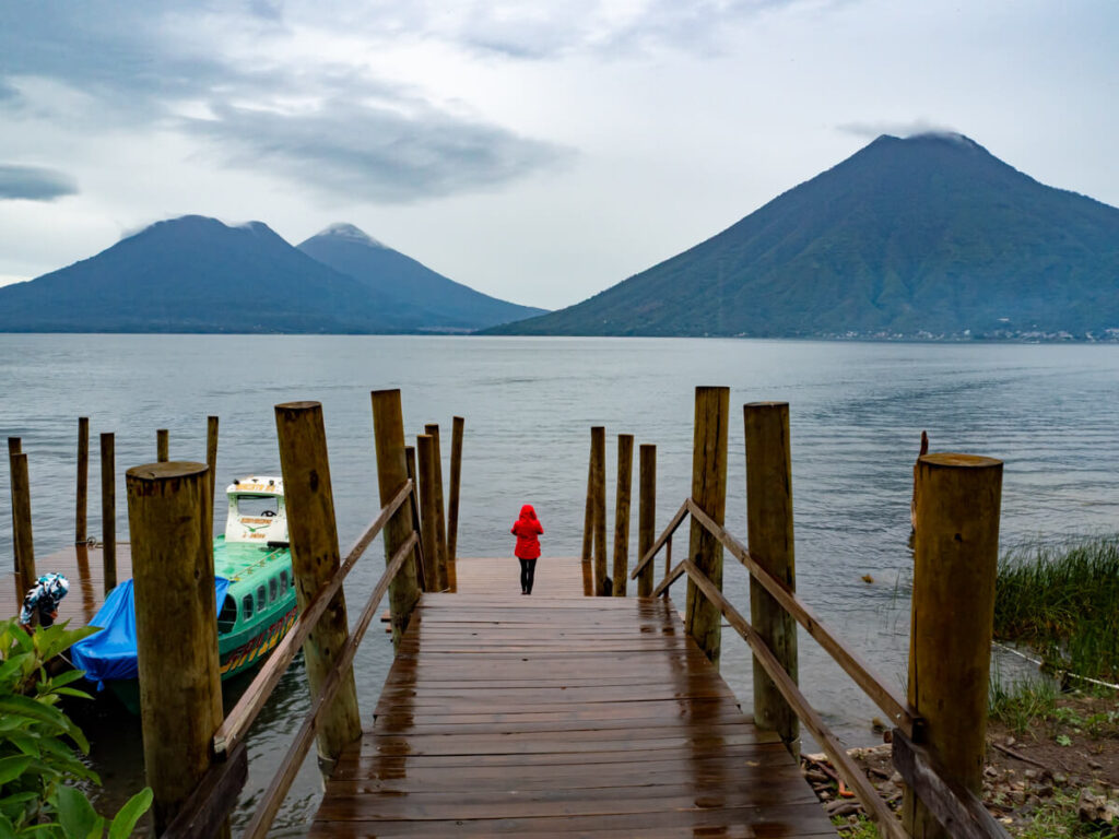 Lucy in a red coat at the end of a wooden dock in San Marcos La Laguna, looking out at three looming volcanoes on the other side of Lake Atitlán.