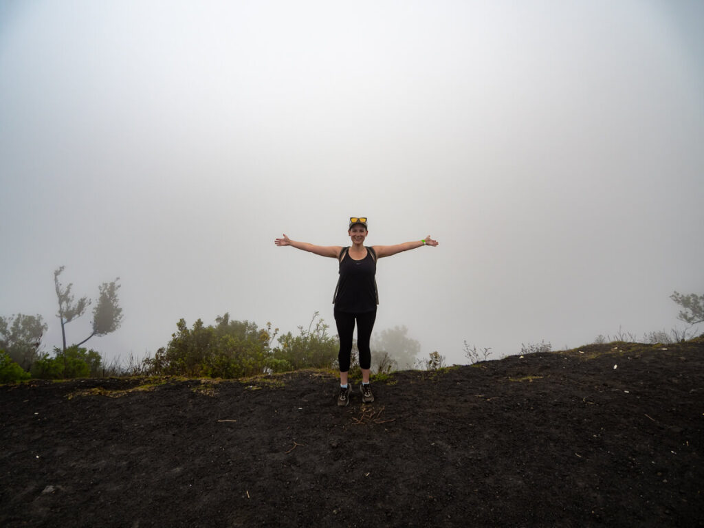 Lucy with her arms wide in front of a blanket of fog on the black lava trails of Pacaya Volcano.