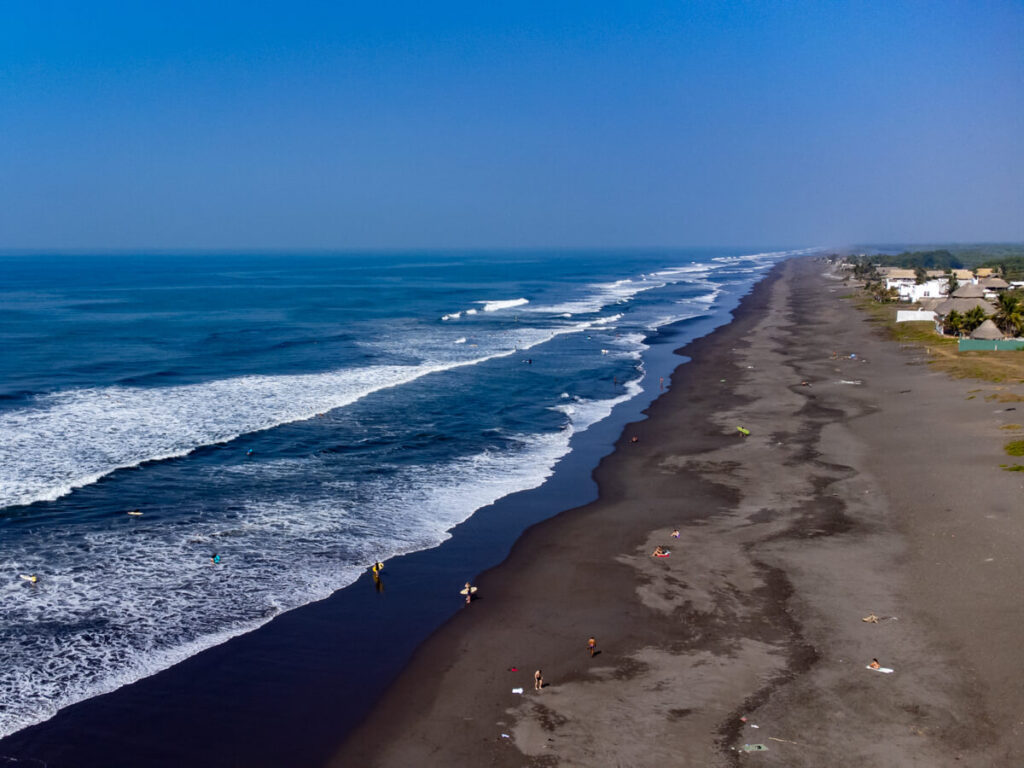 A drone view of a long black sand beach with surfers entering the blue ocean in El Paredón, Guatemala.