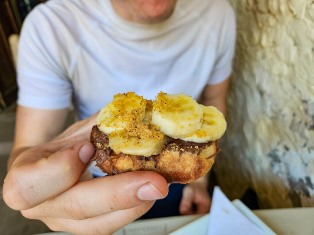 A hand holding coconut bread topped with Nutella and banana.