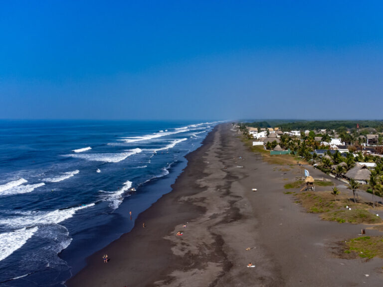 A drone view of a black sand beach reaching for miles, lined by the blue Pacific Ocean and small town of El Paredón, Guatemala.