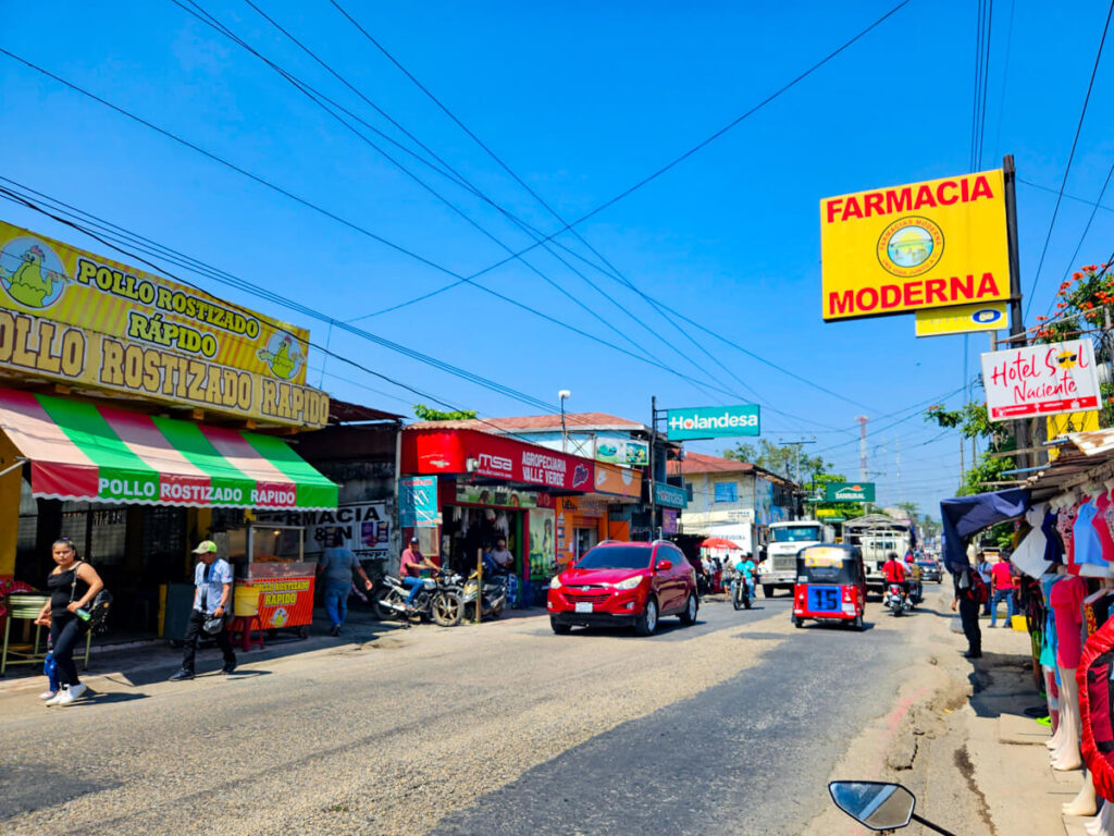 A busy road in Río Dulce town, lined with buildings and being used by pedestrians, cars, and tuk-tuks.