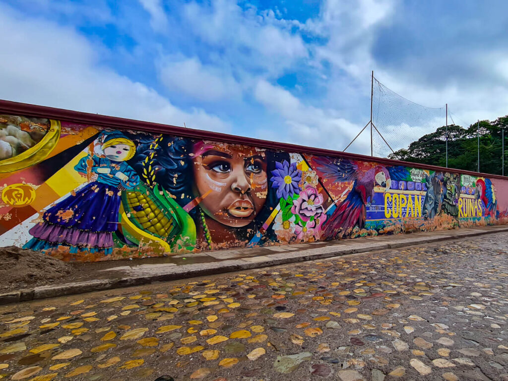 A colourful mural on a wall depicting an indigenous girl, scarlet macaws, and Mayan statues, in Copán Ruinas, Honduras.