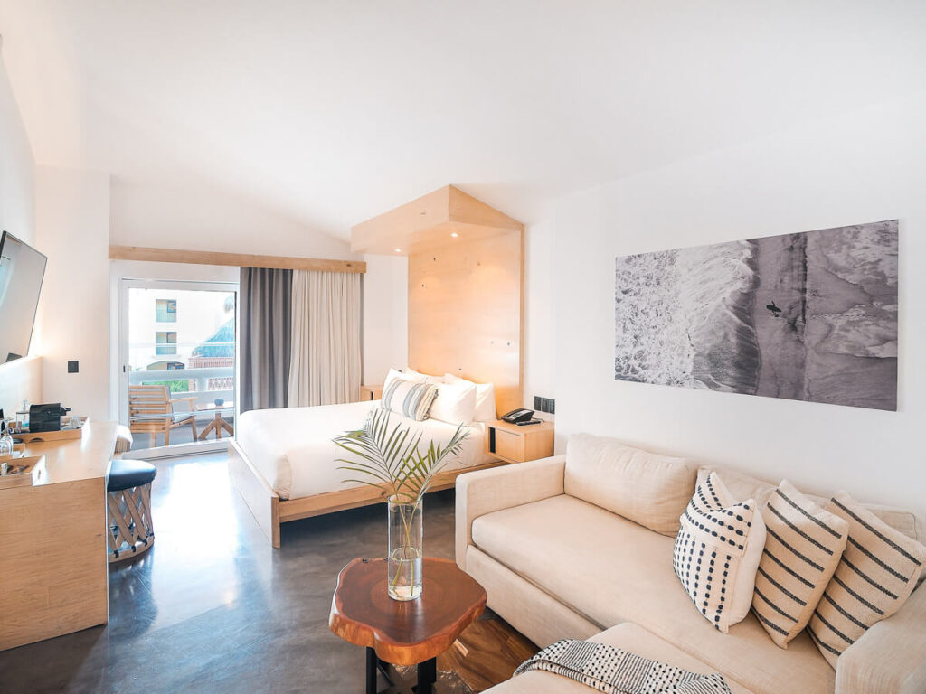 A bright and modern interior of a large hotel room with a plush couch, big bed, and wooden furnishings in Bahia Beach Hotel in Los Cabos.