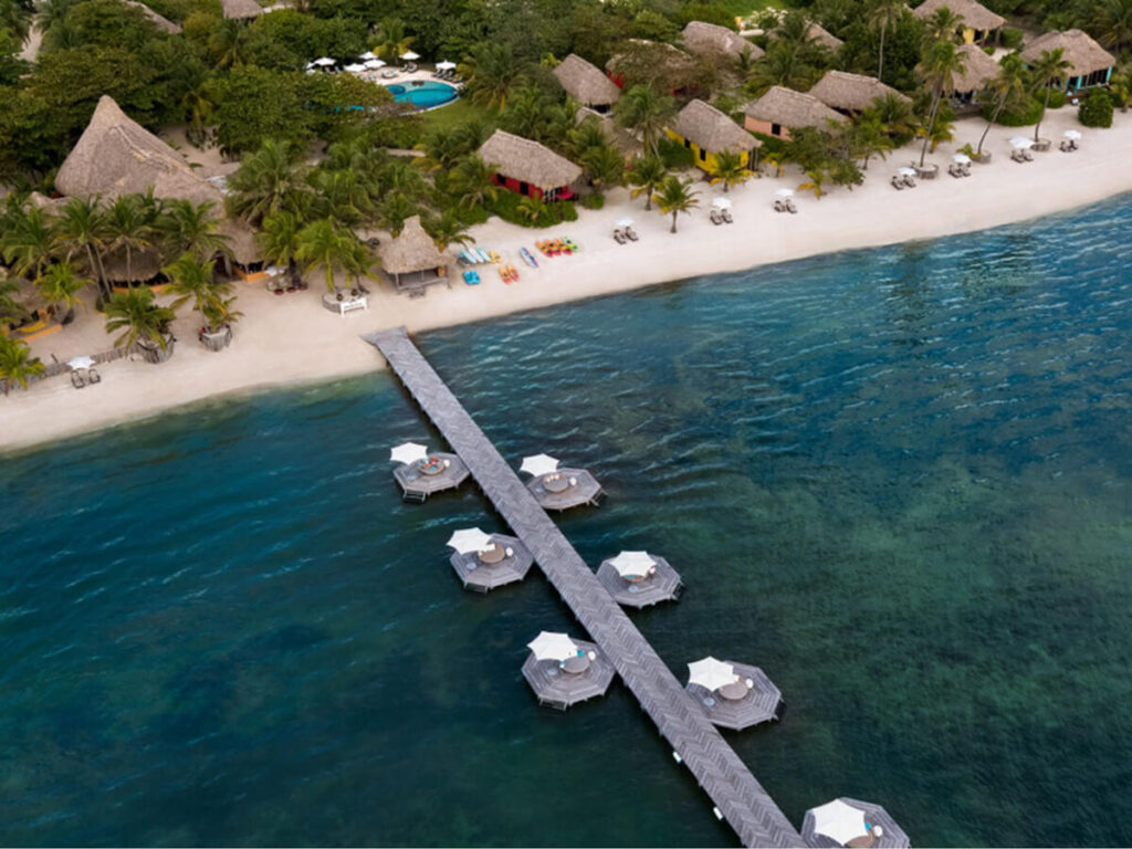 Thatched roofed buildings lining a beach with a long pier on the coast in San Pedro at Matachica Resort and Spa.