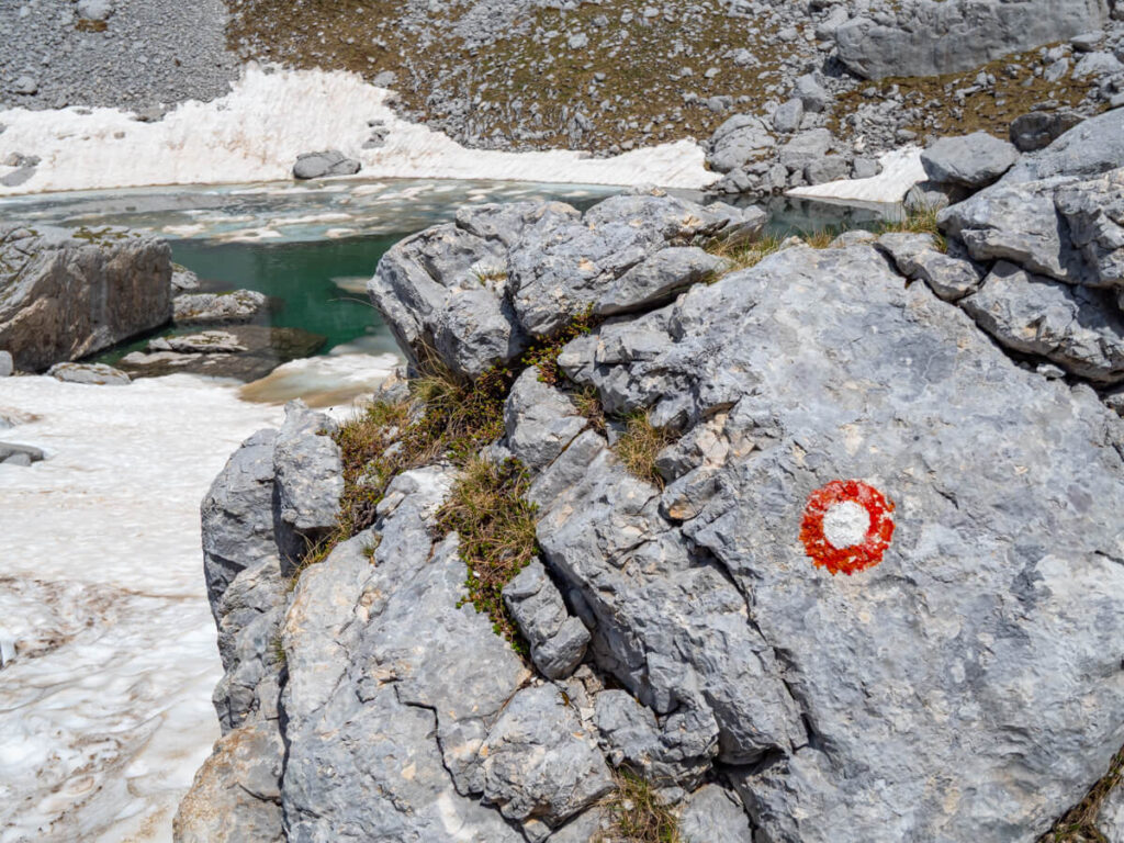 A red and white circular trail marker painted on a boulder in front of the icy blue Zeleni Vir galcial lake on the Bobotov Kuk trail.
