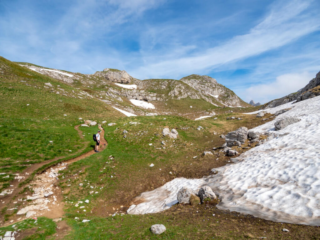 Dan on a dirt trail through a meadow half covered in snow on the way to Bobotov Kuk, one of the best hikes in Durmitor National Park.