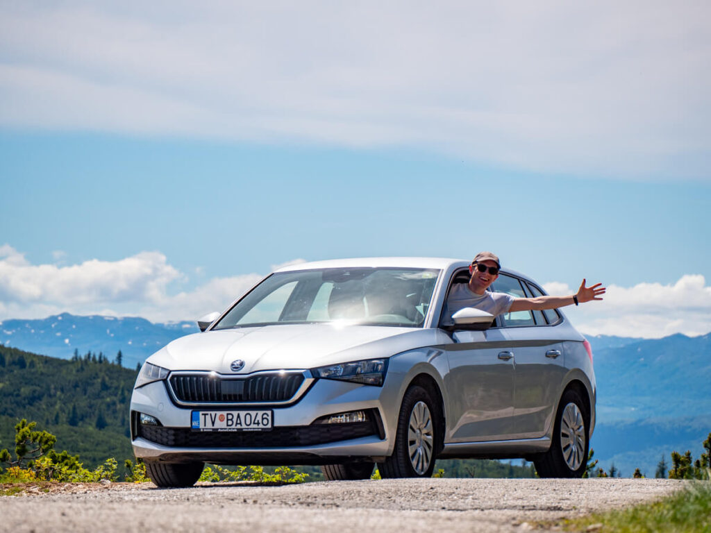Dan hanging out of the driver's window of a silver car with his arm stretched out on a road in Montenegro.