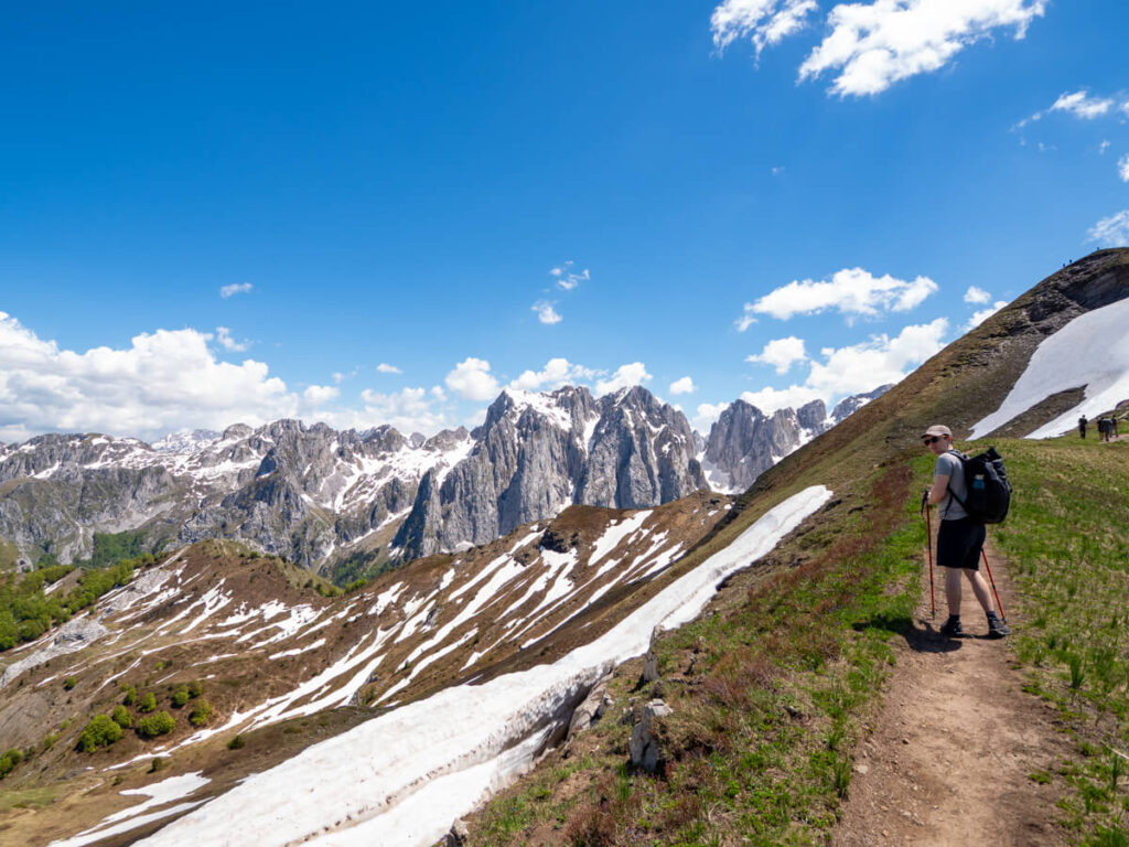 Dan on a dirt trail on a narrow ridge looking out at a valley strewn with snow and the peaks of the Karanfili Massif in the background.