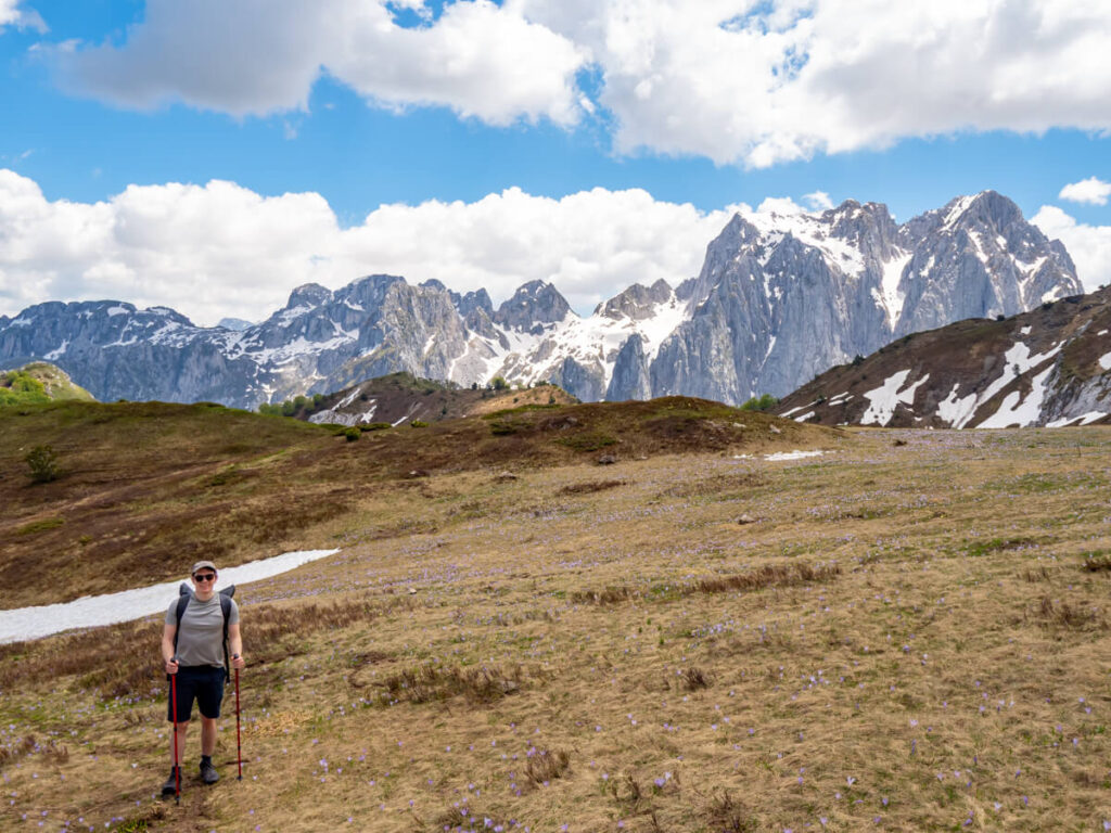Dan hiking in front of a snow field in a valley on the Three Peaks trail in Prokletije National Park. Snowy mountains dominate the backdrop.