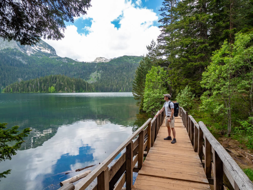 Dan standing on a wooden bridge on the Black Lake trail, looking at the calm glacial surface of the water reflecting the surrounding mountains.