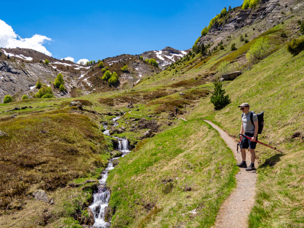 Dan holding trekking poles standing on a path in a meadow looking at a stream on the Three Peaks trail in Prokletije National Park.