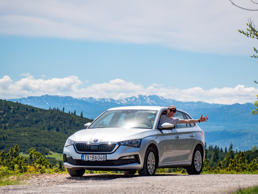 Dan smiling and waving as he leans out of the window of a hire car in Durmitor National Park. The sun is shining and there are mountains and trees in the background.