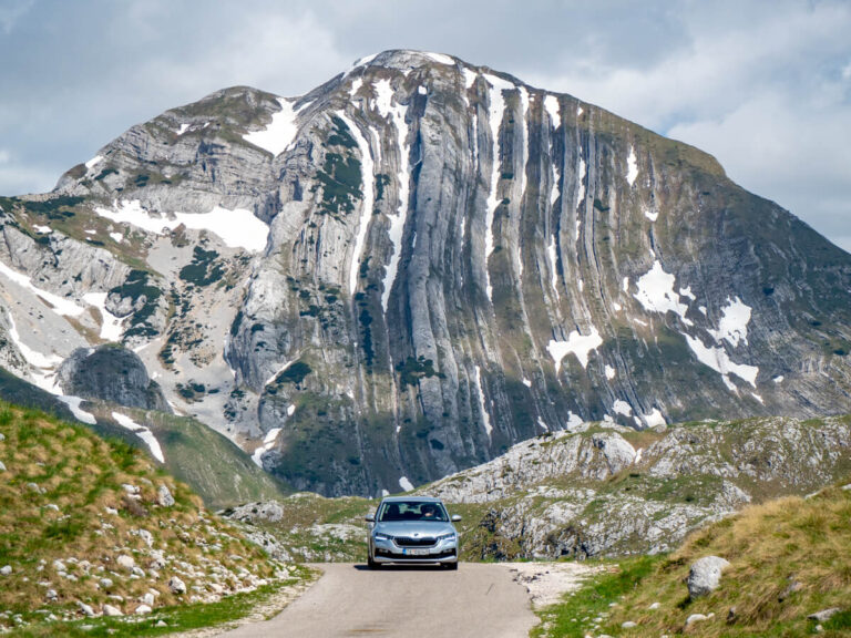 A silver car driving towards the camera, with the looming Prutaš peak in Durmitor National Park in the background.
