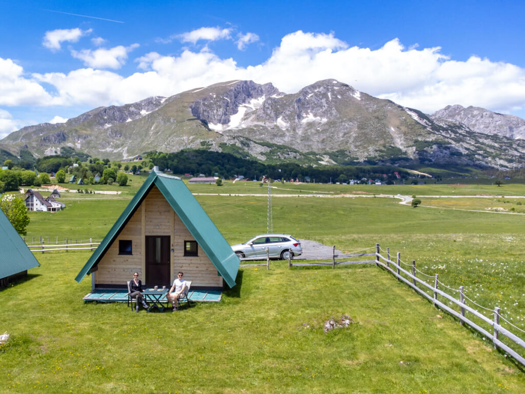 Lucy and Dan sitting outside a wooden A-frame cabin in the sun, backed by the mountains of Durmitor National Park.