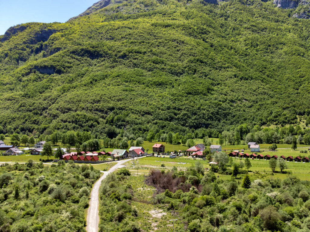 An aerial view of a road leading to buildings under a lush green mountain. Guesthouses including Eko Katun Prokletije are scattered in the Ropojana Valley below.
