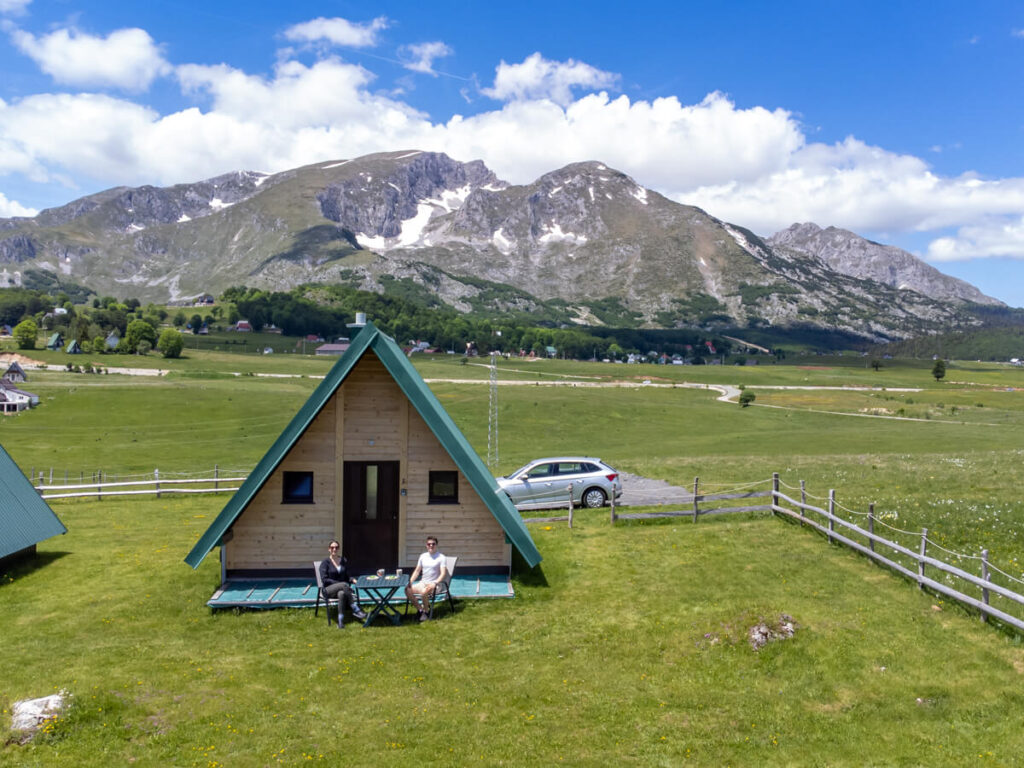 Lucy and Dan sitting outside a wooden A-frame cabin, backed by the mountains of Savin Kuk in Durmitor National Park.