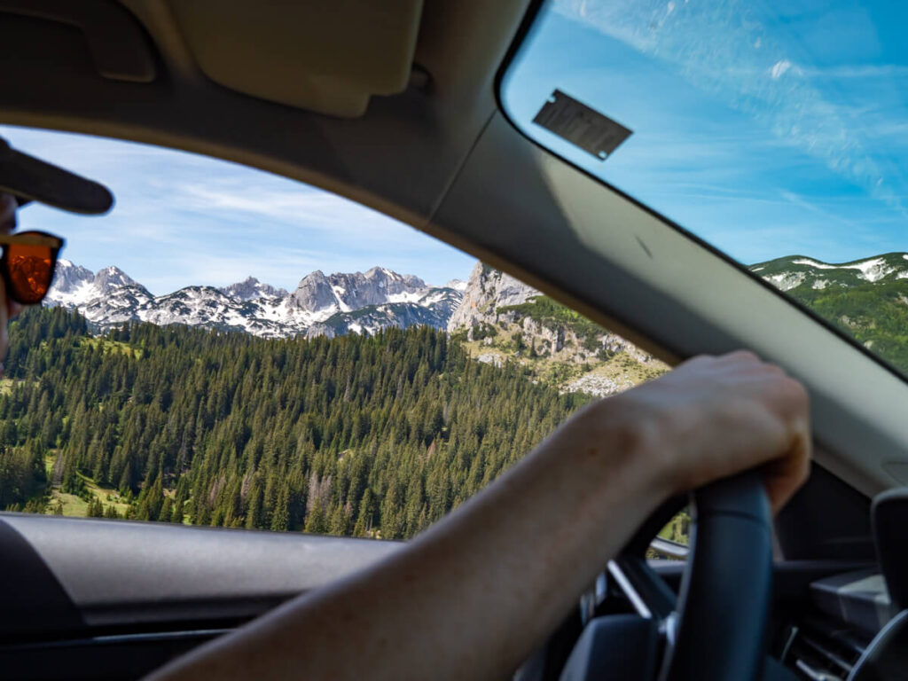 A view of pine forest and snowy peaks out of the driver's window of a car as Dan drives the Durmitor Ring Road.