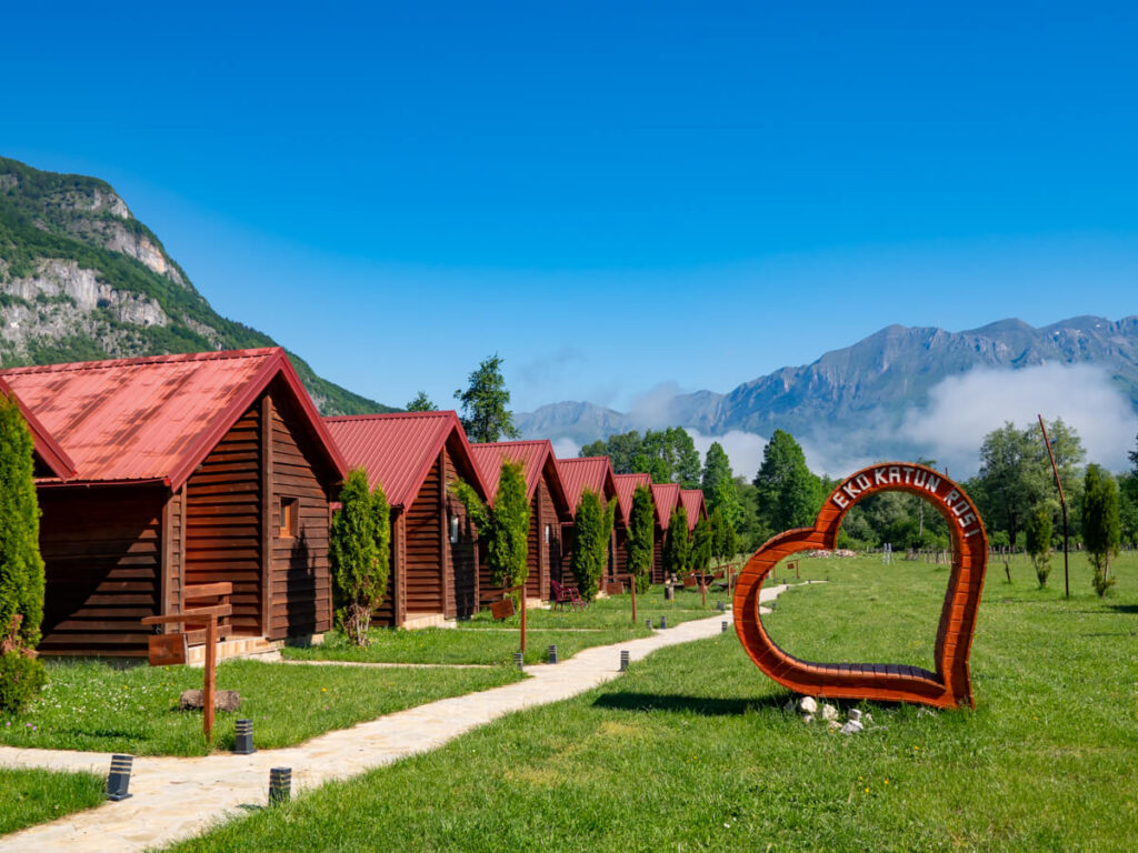A wooden heart sculpture in front of a row of wooden cabins backed by the mountains of the Ropojana Valley at Eko Katun Rosi.